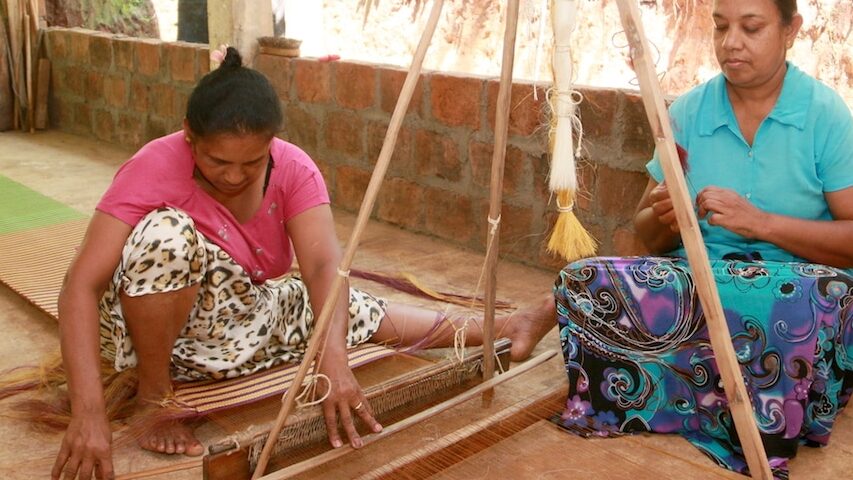 Two women sit on the floor of an open air room weave hana fibers through a traditional loom.