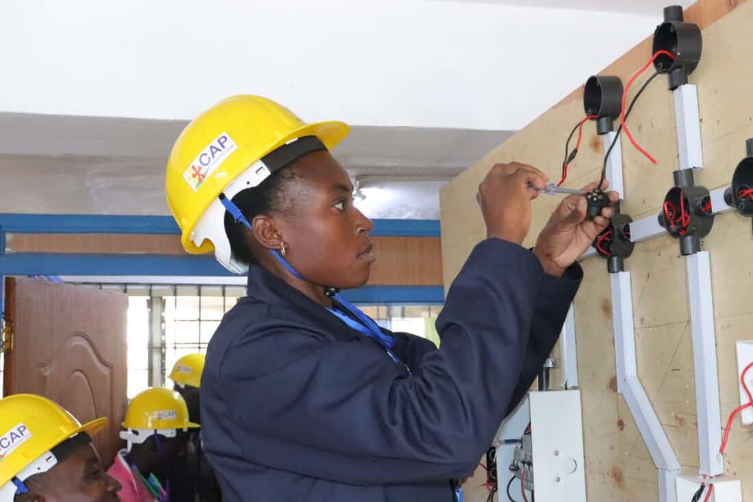 A student wearing a yellow hard hat uses a screwdriver to install electrical wiring on a test wall.